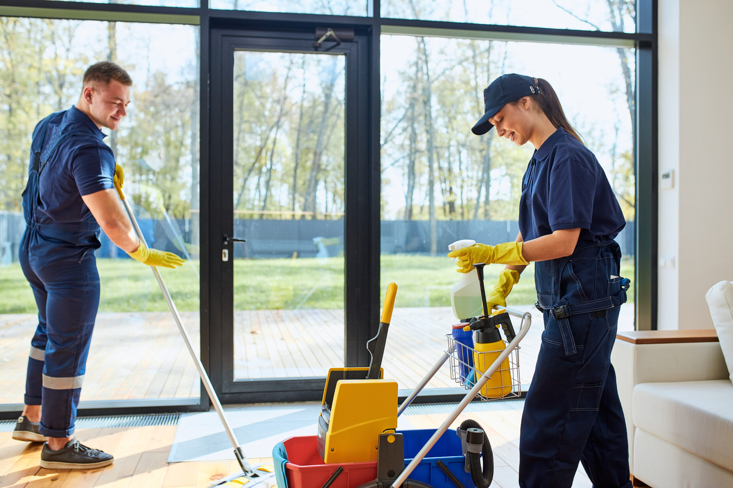 Two professional cleaners in navy uniforms and yellow gloves cleaning a bright living room with large windows, using mops and cleaning supplies