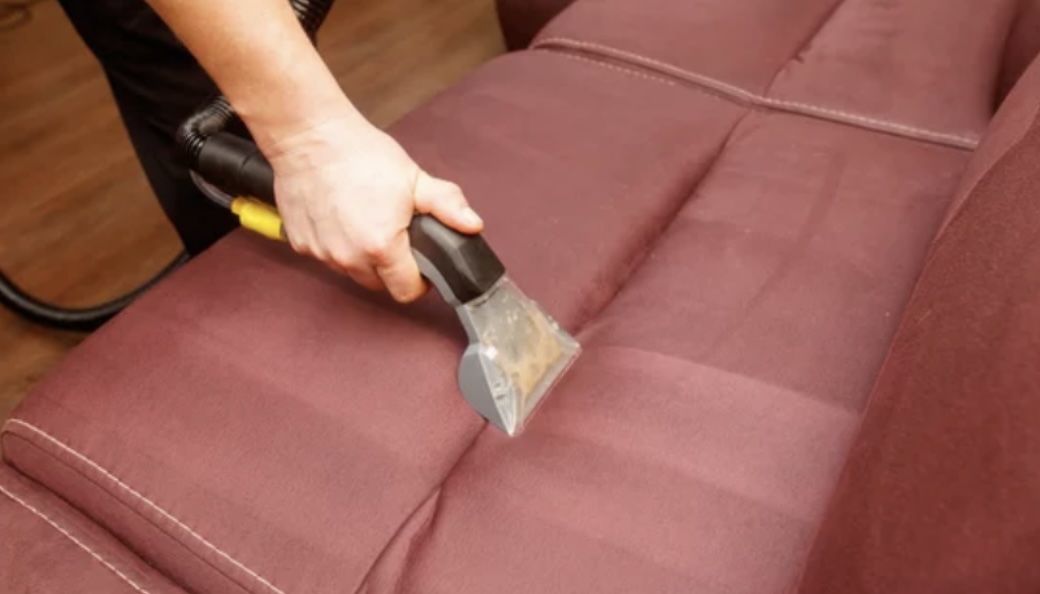 A person using a steam cleaner on a maroon sofa in a room with wooden flooring.